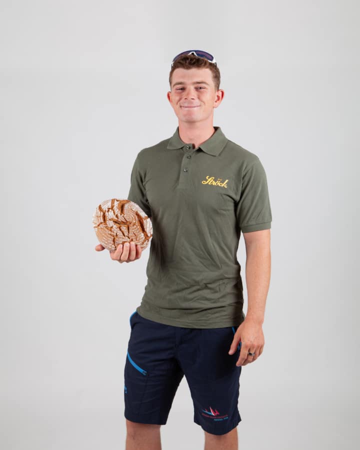 Ein junger Mann in grünem Polohemd und marineblauen Shorts steht vor einem schlichten weißen Hintergrund, lächelt und hält einen runden, in Papier eingewickelten Laib Brot in der rechten Hand. Auf dem Kopf trägt er eine Sonnenbrille.