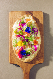 Ein Fladenbrot mit bunten essbaren Blüten, dünn geschnittenen Radieschen, Kräutern und Grünzeug wird auf einem Holzbrett vor einem neutral strukturierten Hintergrund präsentiert.