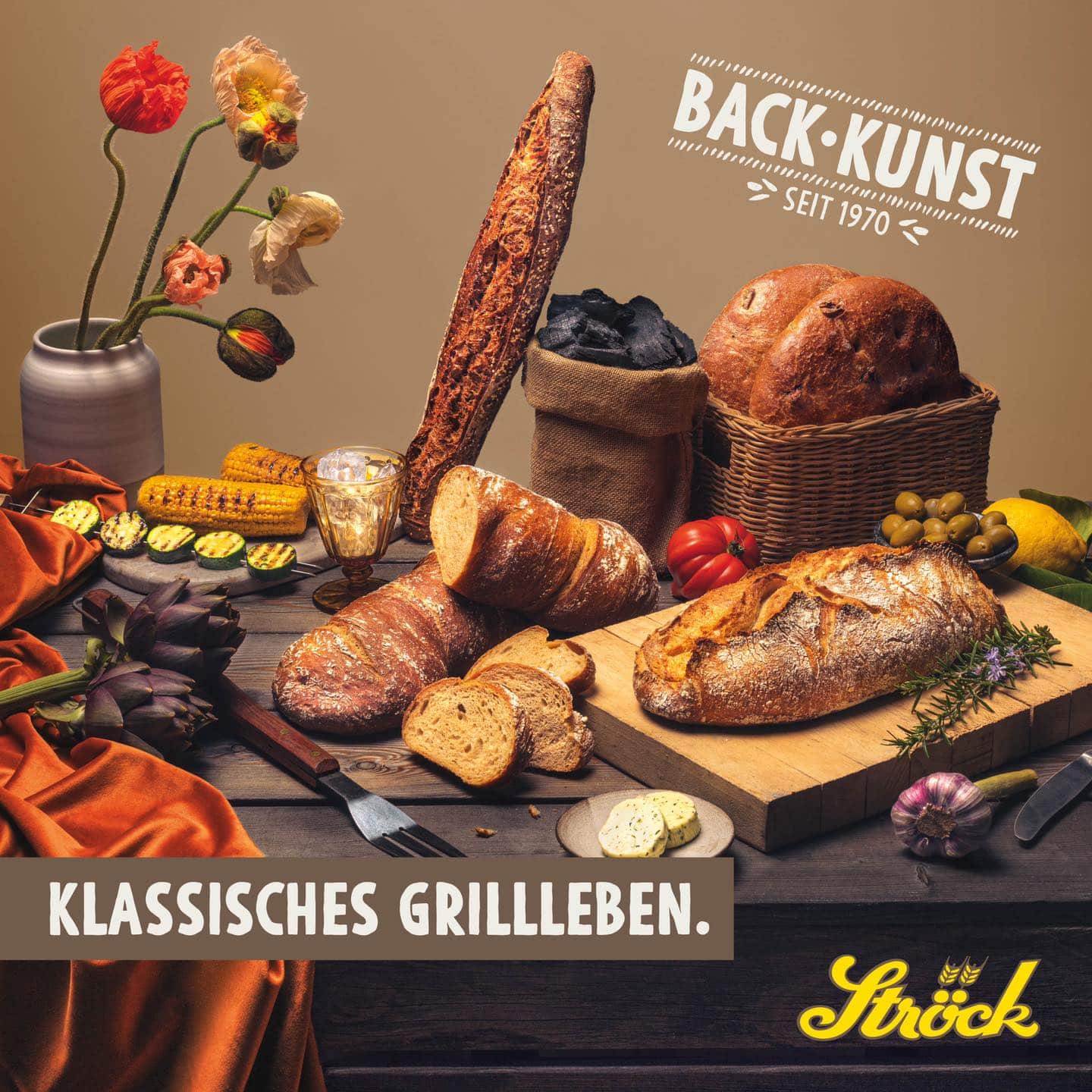 Ein rustikaler Tisch mit verschiedenen Brotsorten, frischem Gemüse, Maiskolben, Käse, einer Blumenvase und einem Brotkorb, mit der Aufschrift "BACK-KUNST SEIT 1970" und "KLASSISCHES GRILLEBEN. Ströck.".