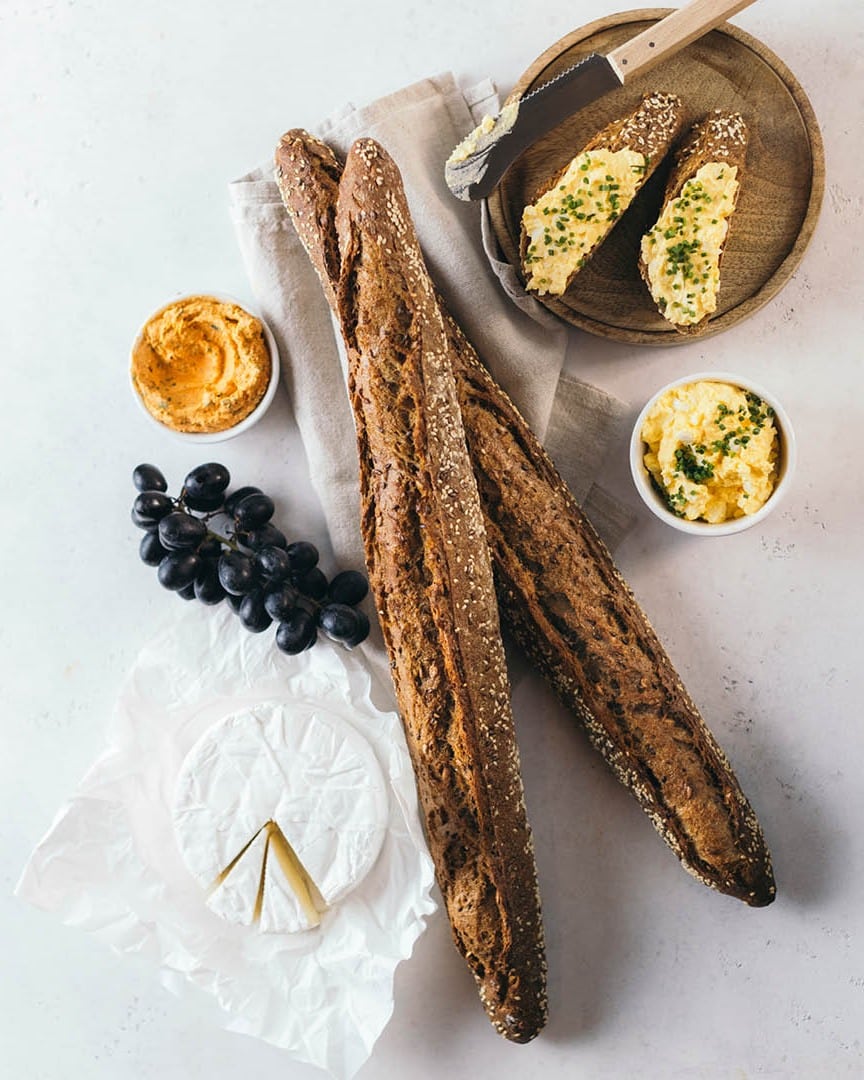 Zwei entkernte Baguettes auf einer Serviette, daneben ein Laib Brie-Käse, eine Traube schwarzer Weintrauben, zwei Stücke Brot mit Aufstrich und Schnittlauch auf einem Holzteller und zwei kleine Schälchen mit Aufstrichen auf einer weißen Fläche.