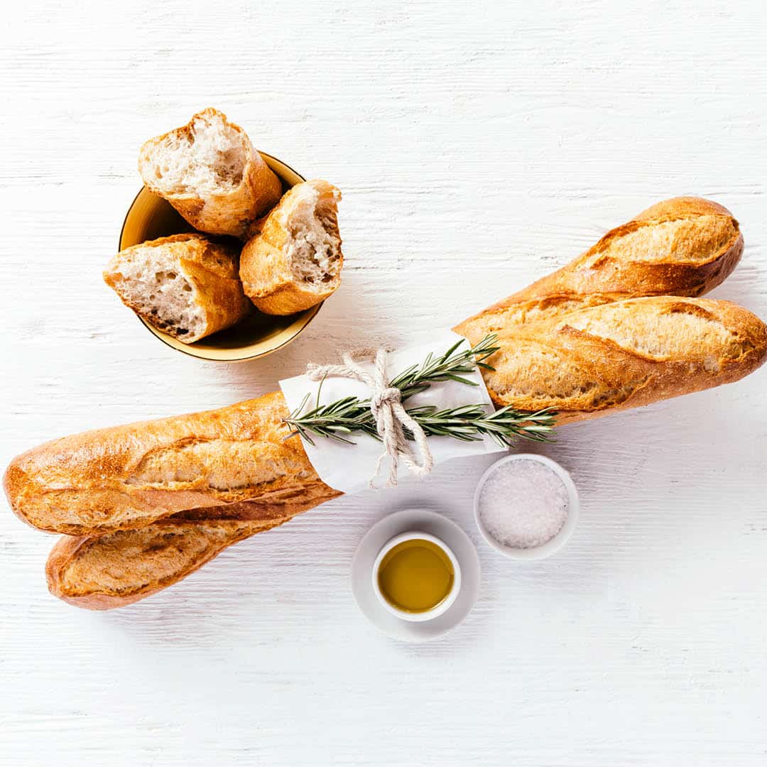 Zwei mit Rosmarin gebundene Baguettes, eine Schale mit Brotstücken und kleine Schalen mit Olivenöl und Salz sind auf einer weißen Fläche angeordnet.
