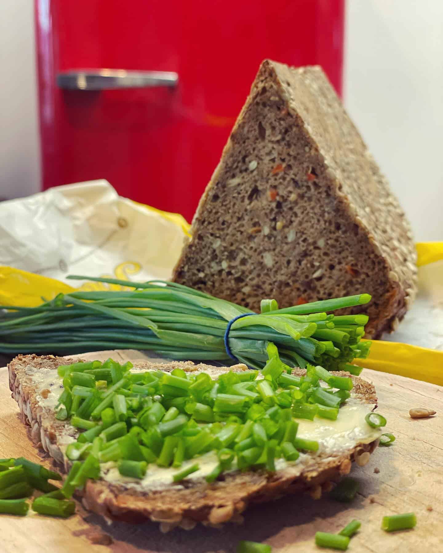 Auf einem Schneidebrett liegt eine Scheibe Vollkornbrot mit Butter und gehacktem Schnittlauch. Dahinter liegen ein Bündel frischer Schnittlauch und ein großer, dreieckiger Laib Körnerbrot, mit einem roten Kühlschrank im Hintergrund.