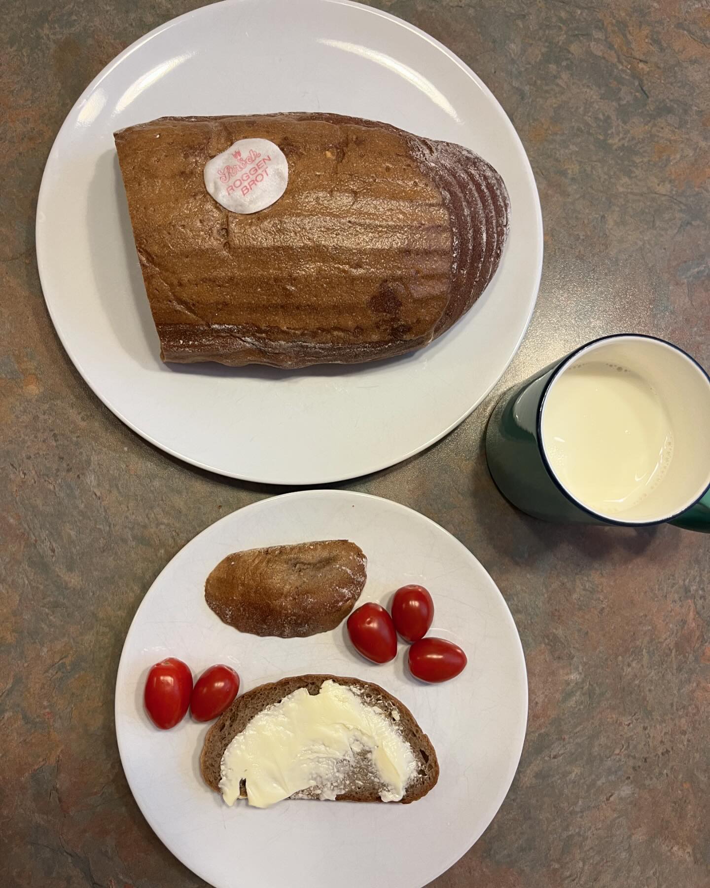 Zwei Teller: auf dem einen liegt ein Brot mit der Aufschrift Weizenbrot, auf dem anderen liegen Brotscheiben - eine mit Butter, eine ohne - neben Kirschtomaten. Rechts daneben steht ein grüner Milchbecher auf einer braunen, marmorierten Fläche.