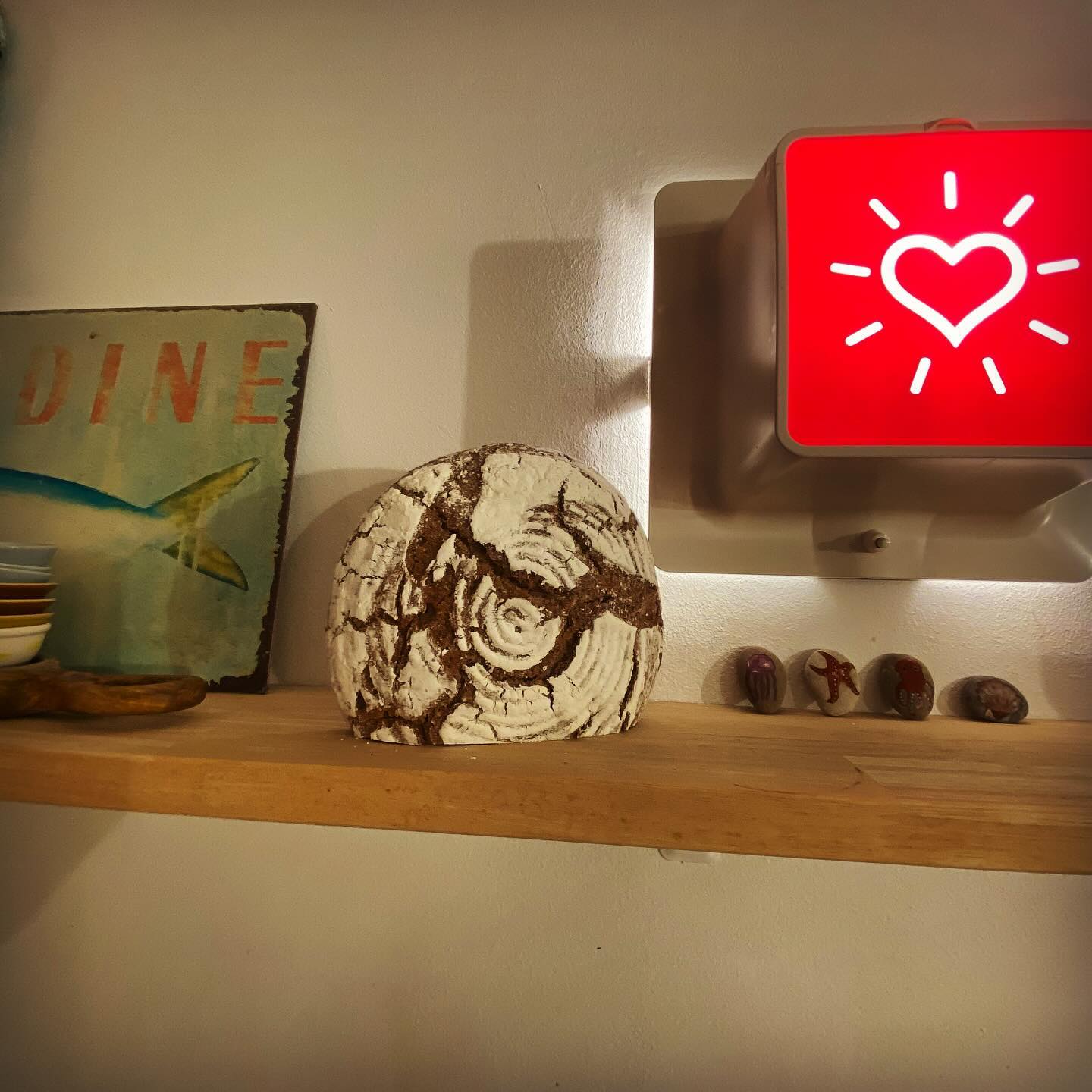 Auf einem Holzregal stehen ein fossilartiger Stein, ein Retro-DINE"-Schild mit einem Fisch, ein Stapel kleiner Schalen, vier bemalte Steine und ein roter Leuchtkasten mit einem leuchtenden weißen Herzsymbol.