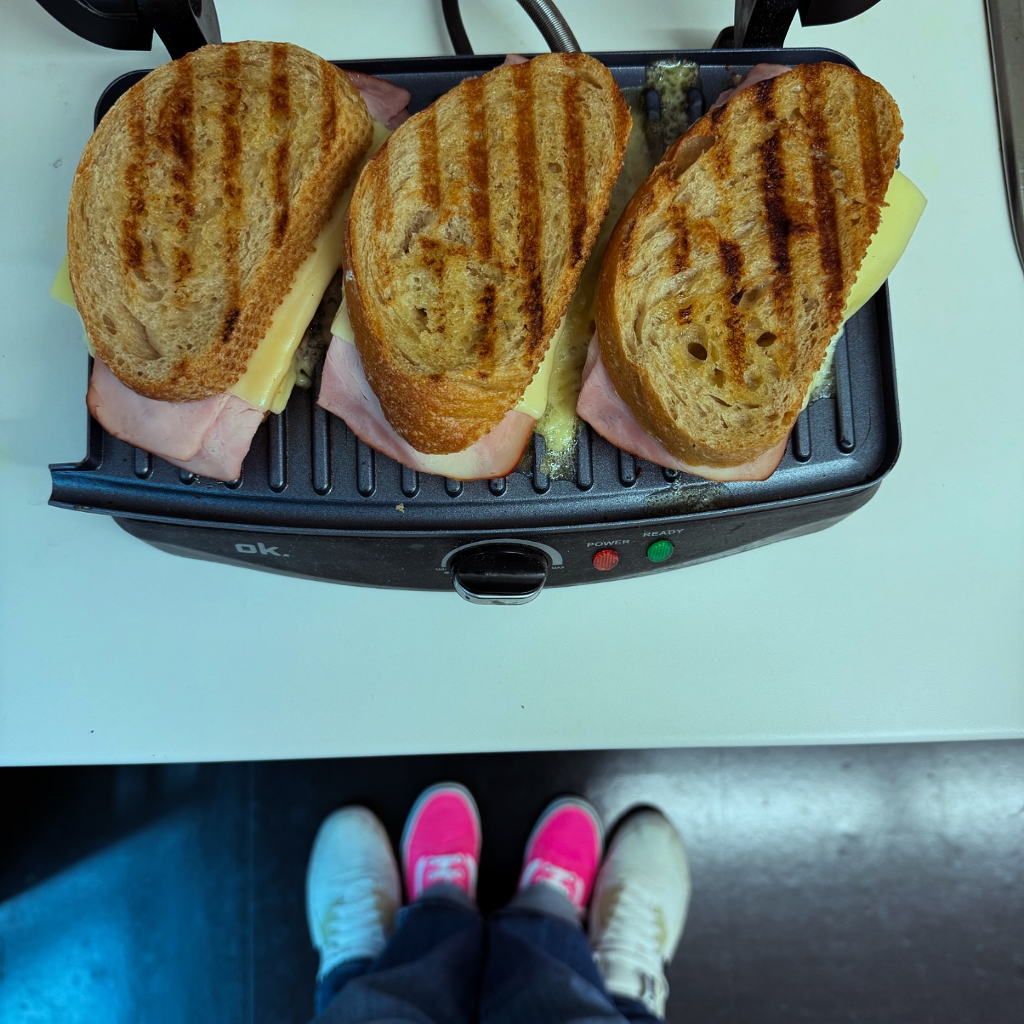Drei Sandwiches mit gegrilltem Brot, Schinken und Käse liegen in einer Panini-Presse. Unter der Theke steht eine Person in weißen und neonpinken Turnschuhen auf einem dunklen Boden.