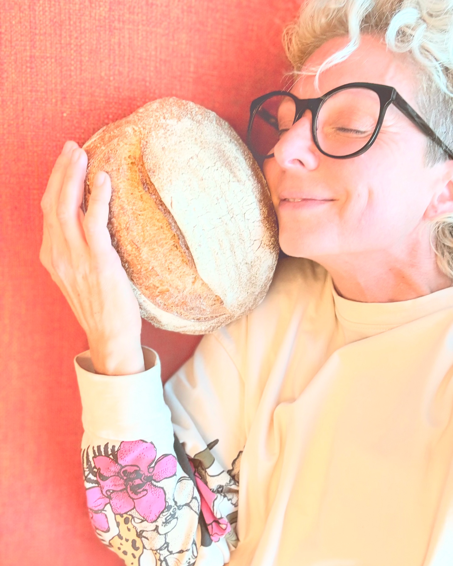 Eine Person mit kurzen, hellen Haaren und schwarzer Brille drückt sich fröhlich einen runden Laib Brot an die Wange. Sie trägt ein helles Hemd mit rosafarbenen Blumenmustern, und der Hintergrund ist in einem warmen Korallton gehalten.