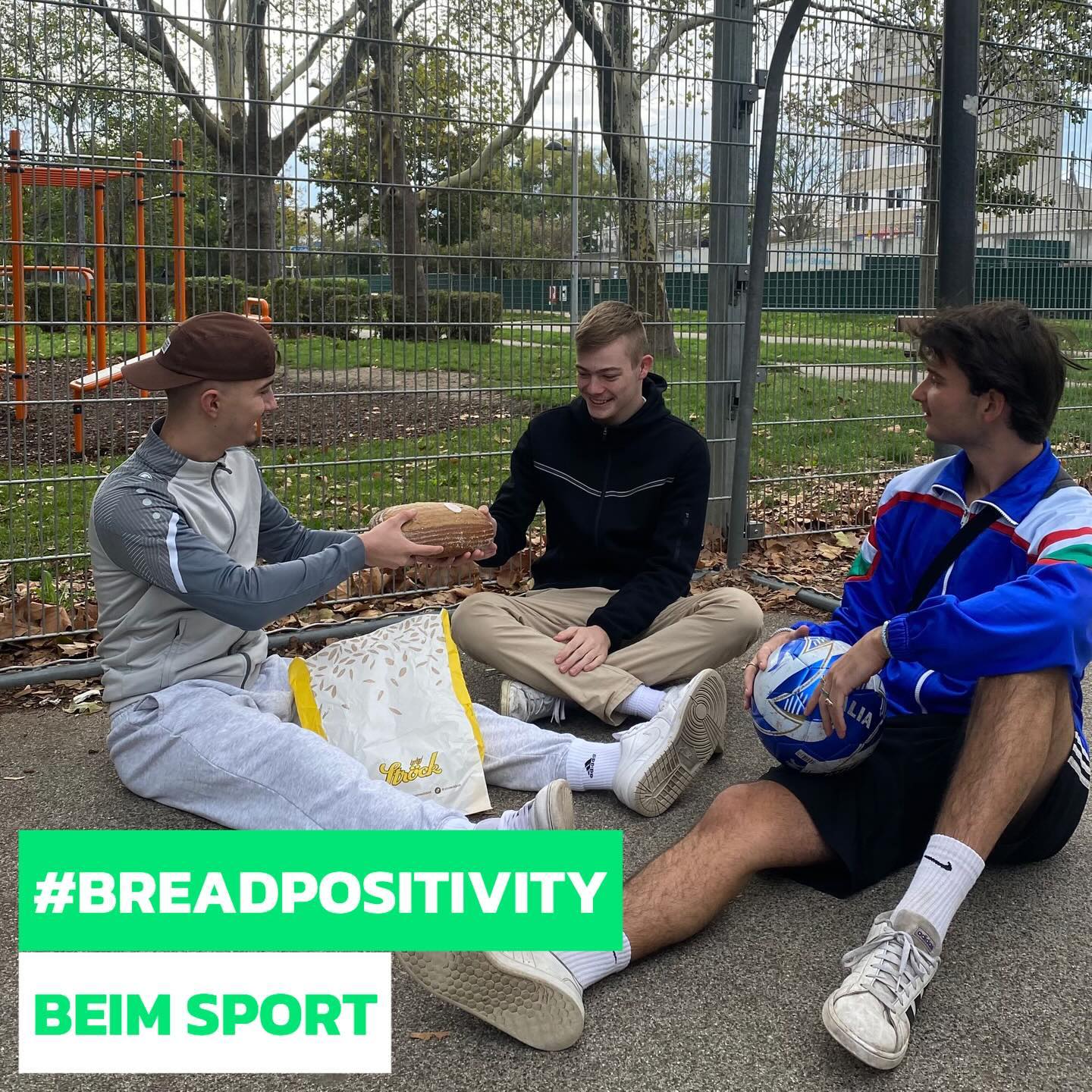 Drei junge Männer sitzen auf dem Boden an einem Zaun in einem Park. Einer reicht einem anderen einen Laib Brot, während der dritte einen Fußball hält. Der Text #BREADPOSITIVITY BEIM SPORT erscheint am unteren Rand.