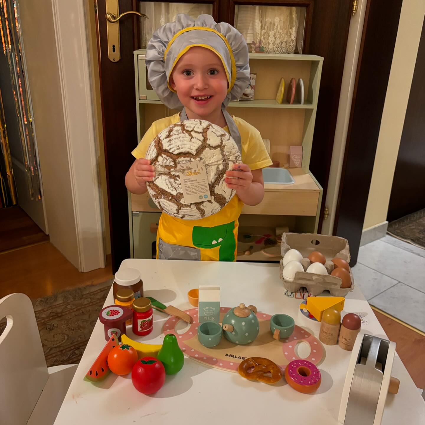 Ein lächelndes kleines Kind mit Kochmütze und Schürze hält einen großen runden Laib Brot in der Hand. Es steht hinter einem Spielküchen-Set mit Spielzeug-Essen, einer Teekanne, Tassen und verschiedenen vorgetäuschten Lebensmitteln auf einem kleinen weißen Tisch.