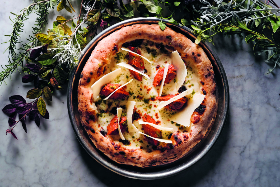 Eine im Holzofen gebackene Pizza mit Tomatensoße, geschmolzenem Käse und großen Parmesanspänen liegt auf einem Metalltablett, umgeben von frischen grünen und violetten Kräutern auf einer Marmorfläche.