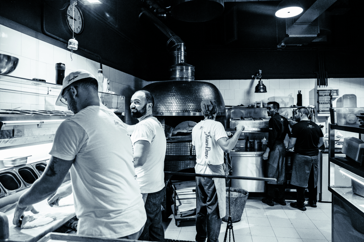Eine belebte Restaurantküche, in der mehrere Köche arbeiten, von denen einige an der Theke und andere in der Nähe des Ofens Speisen zubereiten. Die Szene ist in Schwarz-Weiß gehalten und vermittelt eine lebendige und doch klassische Atmosphäre.