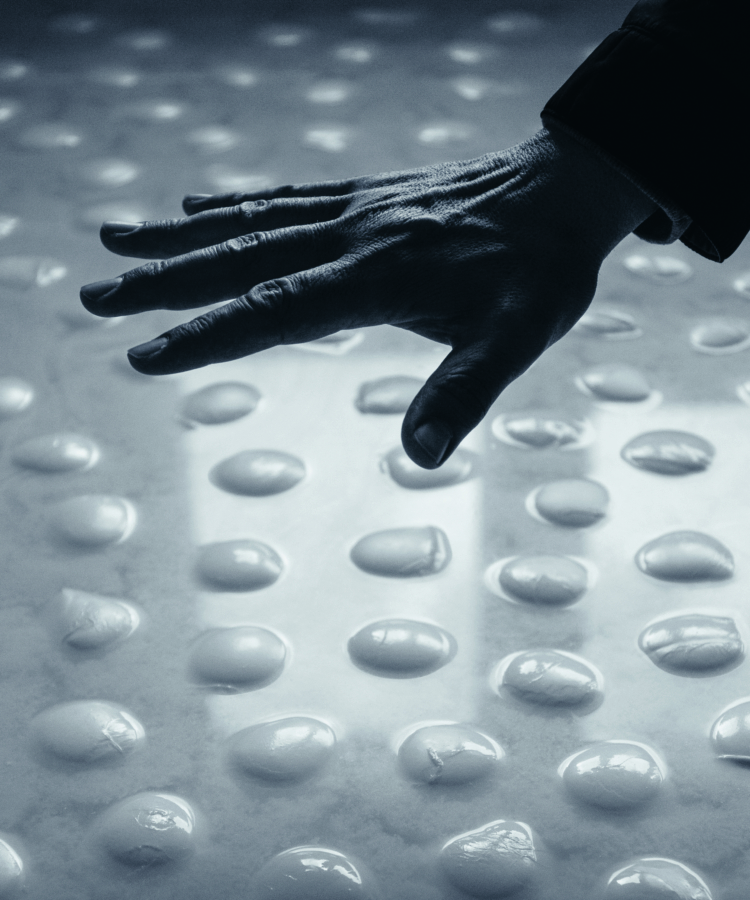 Eine Hand schwebt über einer Oberfläche, die mit gleichmäßig verteilten, glänzenden, abgerundeten Unebenheiten bedeckt ist. Das Bild ist in kühlen Blautönen gehalten, die eine reflektierende, abstrakte und taktile Atmosphäre schaffen.