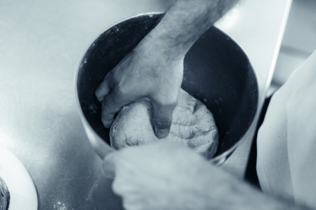 Eine Person knetet Teig mit der Hand in einer Metallschüssel auf einem Küchentisch und bereitet ihn zum Backen vor. Das Bild ist in schwarz-weiß gehalten.