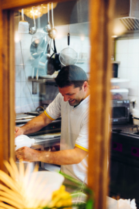 Ein Mann in einem weißen Hemd lächelt, während er in einer Küche, umgeben von hängenden Töpfen und Utensilien, Zutaten mit einem Standmixer vermischt, gesehen durch ein Fenster mit Holzrahmen.