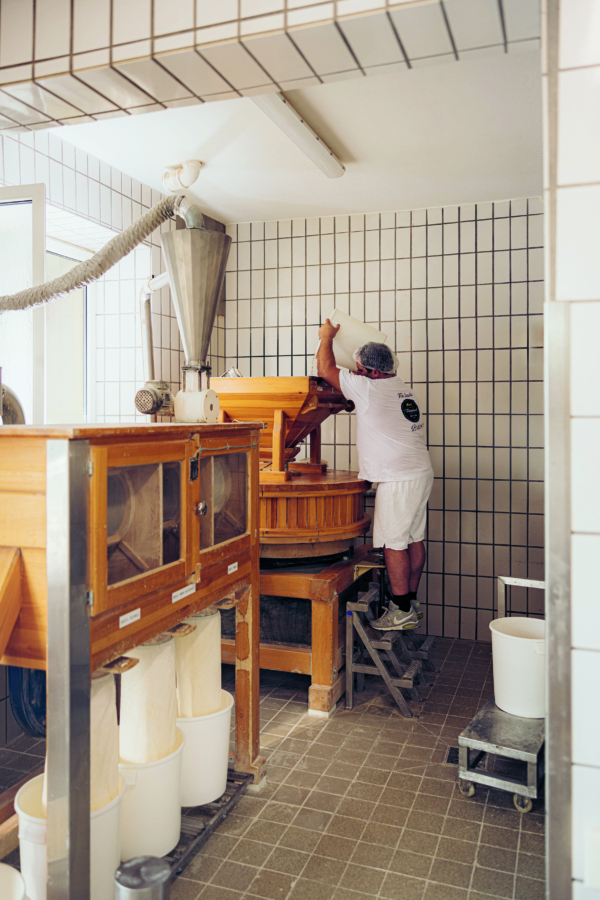 Eine Person in weißer Arbeitskleidung steht auf einem Trittschemel und schüttet Zutaten in eine große hölzerne Fräsmaschine in einem gefliesten, industriellen Raum für die Lebensmittelverarbeitung. Mehrere Behälter und Geräte umgeben den Arbeitsbereich.