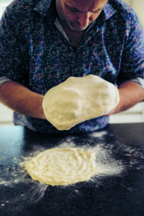 Eine Person formt mit beiden Händen Teig über einer bemehlten Fläche auf einer dunklen Arbeitsplatte und bereitet sich auf die Zubereitung von Pizza oder Brot vor. Die Person blickt nach unten und trägt ein gemustertes Hemd.