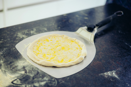 Eine rohe, mit Öl bestrichene Pizzakruste liegt auf einem metallenen Pizzaschieber auf einer bemehlten, dunklen Arbeitsplatte und ist bereit zum Backen.