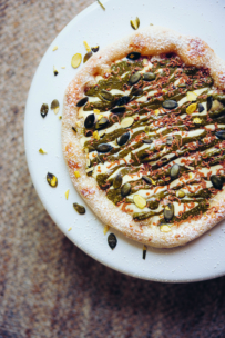Eine runde Pizza, belegt mit grünem Pesto, Kürbiskernen und einem leichten Spritzer Sauce, liegt auf einem weißen Teller vor einem strukturierten beigen Hintergrund.