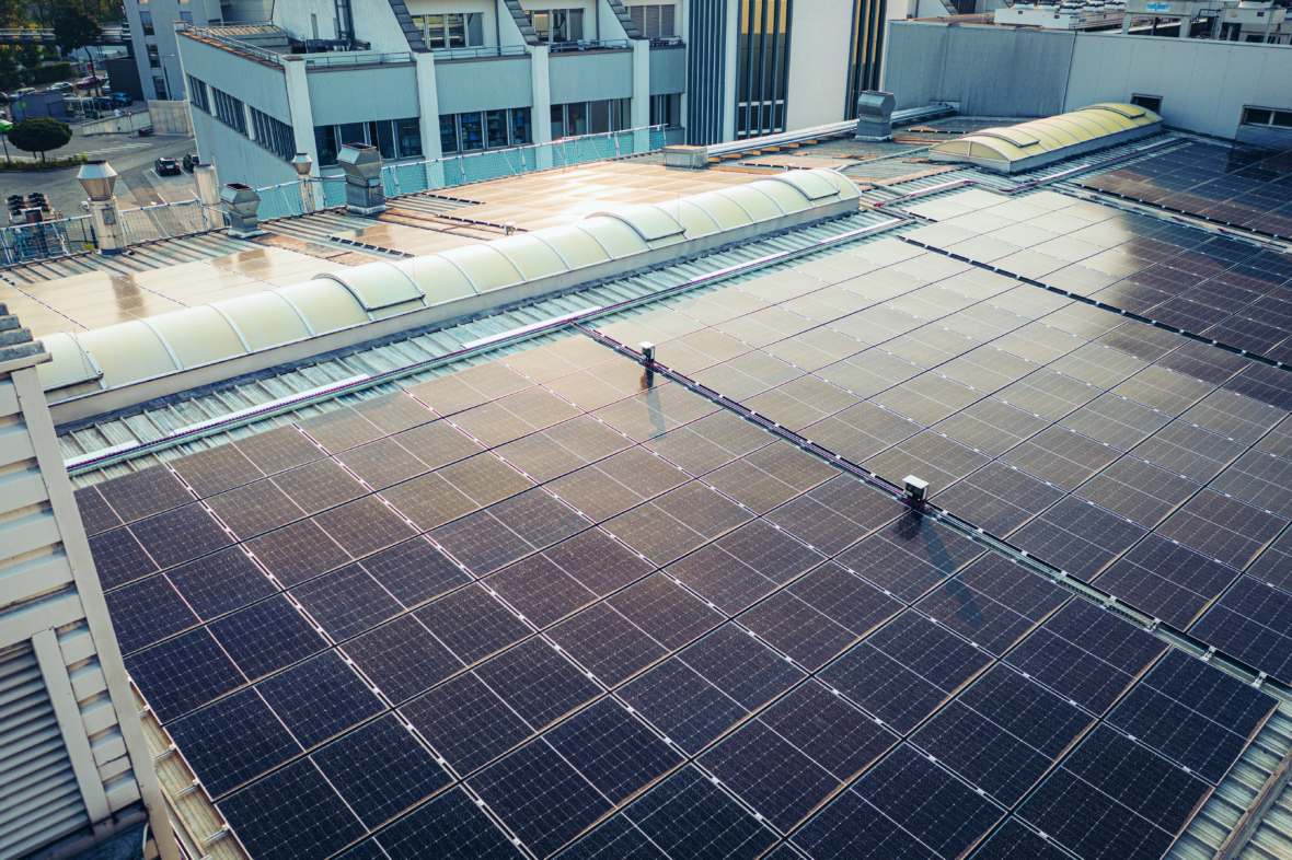 Auf dem Dach eines modernen Industriegebäudes ist eine große Anordnung von Solarmodulen installiert, die das Sonnenlicht zur Erzeugung erneuerbarer Energie einfängt, während im Hintergrund nahe gelegene Gebäude zu sehen sind.