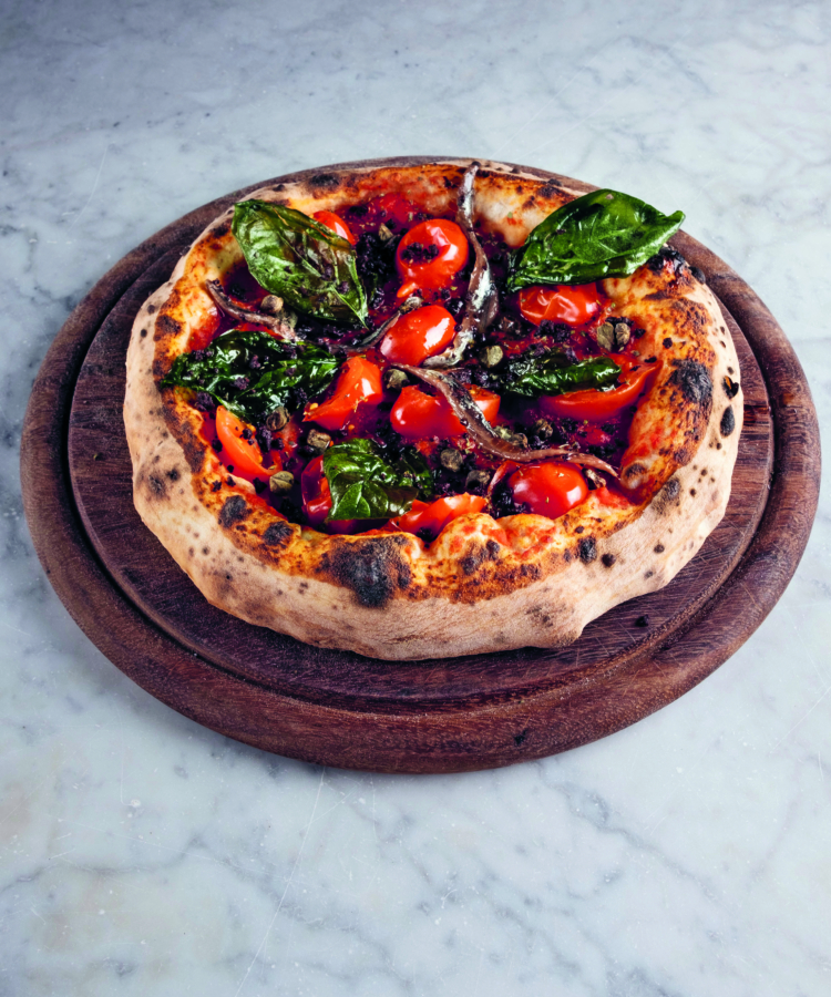 Eine im Holzofen gebackene Pizza, belegt mit Kirschtomaten, Basilikum, Oliven und Sardellen, liegt auf einem runden Holzbrett über einer Marmorplatte.