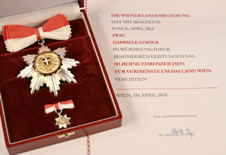 Nahaufnahme eines Auszeichnungssets in einer roten Schachtel mit einer Gold- und Silbermedaille mit rot-weißem Band neben einer Urkunde zur Ehrung von Gabriele Ströck für besondere Leistungen in Wien, datiert April 2024.