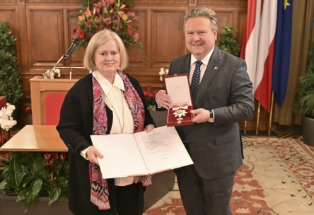 Eine Frau hält eine offene Urkunde in der Hand, während ein Mann im Anzug lächelnd neben ihr steht und eine Medaille in einer roten Schachtel zeigt. Sie befinden sich in einem Innenraum, mit Blumen, Pflanzen und Fahnen im Hintergrund.