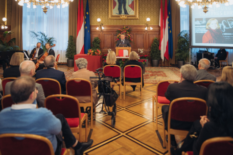 Ein Redner spricht zu einem Publikum in einem formellen, verzierten Raum mit roten Stühlen, Kronleuchtern und Fahnen. Auf der linken Seite spielt ein Musikertrio, und auf der rechten Seite zeigt ein Projektor das Bild einer Frau auf einer Leinwand.