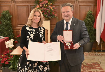 Eine Frau in einem schwarzen Kleid mit weißen Blumenmustern hält eine offene Urkunde, während ein Mann im Anzug neben ihr steht und eine verzierte Medaille in einer roten Schachtel hält. Sie stehen in einem eleganten Raum mit Holzvertäfelung und Blumenarrangements.