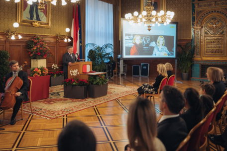 Ein Mann spricht an einem Podium in einem prunkvollen Raum mit Holzverkleidung und Kronleuchtern. Die Menschen sitzen ihm gegenüber, und auf einem großen Bildschirm wird eine Präsentation mit zwei Frauen und Text in polnischer Sprache gezeigt. Blumen und ein Cellist sind ebenfalls anwesend.