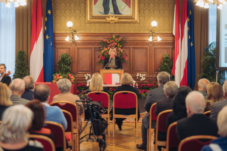 Eine Frau spricht auf einem Podium in einem mit roten und weißen Fahnen geschmückten Raum vor einem Publikum, das auf roten Stühlen sitzt. Blumenarrangements und gerahmte Kunstwerke schmücken den Raum und verleihen ihm eine formelle, offizielle Atmosphäre.