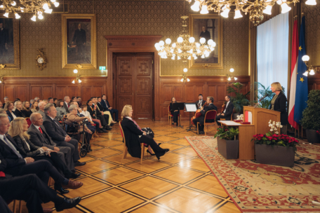 Eine Frau spricht an einem Podium in einem prunkvollen Raum mit Kronleuchtern, großen Porträts und einem Publikum, das ihr gegenüber sitzt. Musiker sitzen in der Nähe und zwei Fahnen, darunter die EU-Flagge, stehen am Fenster.