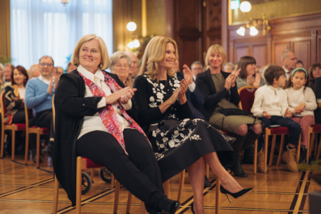 Zwei Frauen sitzen bei einer feierlichen Veranstaltung in der ersten Reihe, lächeln und klatschen. Hinter ihnen applaudiert ein Publikum aus Erwachsenen und Kindern. Die Kulisse ist elegant, mit verzierten Holztafeln und warmer Beleuchtung.