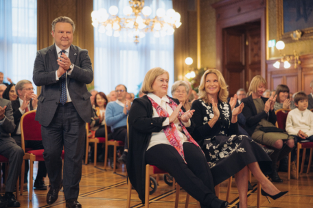 Ein Mann im Anzug steht auf und klatscht, während zwei Frauen in der ersten Reihe ebenfalls applaudieren. Das Publikum hinter ihnen sitzt auf roten Stühlen in einem großen, eleganten Raum mit Kronleuchtern und kunstvollem Dekor.