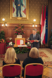 Ein Mann im Anzug steht an einem Podium und liest in einem mit Fahnen, Blumen und einem großen Gemälde geschmückten Raum aus einer Zeitung vor. Zwei Frauen sitzen ihm gegenüber. Die Atmosphäre ist formell und offiziell.