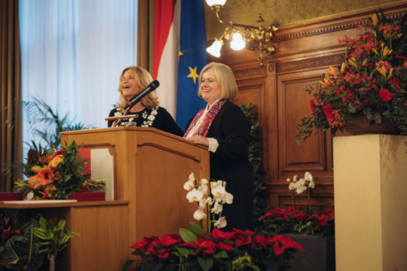 Zwei Frauen stehen lächelnd an einem blumengeschmückten Holzpodium in einem eleganten Raum, mit österreichischen und EU-Flaggen und einer kunstvollen Holzvertäfelung im Hintergrund. Die Szenerie deutet auf eine formelle oder offizielle Veranstaltung hin.