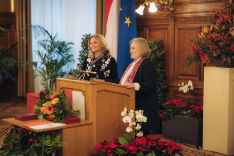 Zwei Frauen stehen an einem hölzernen Podium in einem formellen Raum, der mit roten Blumen und Pflanzen geschmückt ist. Hinter ihnen sind die Flaggen Österreichs und der Europäischen Union zu sehen. Eine Frau spricht, beide scheinen in die Veranstaltung vertieft zu sein.