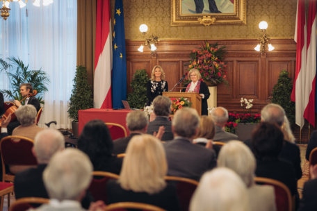 Zwei Frauen stehen an einem Podium und sprechen zu einem Publikum in einem formellen, geschmückten Raum mit Fahnen, Blumen und einem sitzenden Musiker auf der linken Seite. Das Publikum sitzt mit Blick auf die Redner.