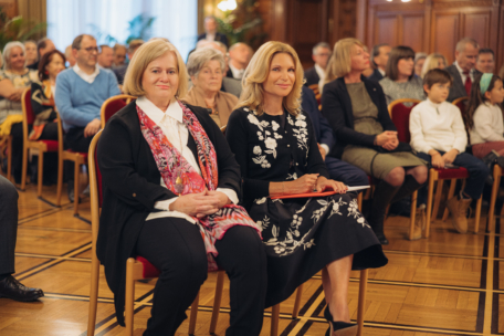 Zwei Frauen sitzen lächelnd in der ersten Reihe einer Indoor-Veranstaltung, hinter ihnen sitzen Erwachsene und Kinder in einem formellen Rahmen mit holzgetäfelten Wänden und polierten Böden.