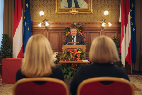Ein Mann steht an einem Podium und hält eine Rede in einem verzierten Raum mit Holzvertäfelung und Fahnen, während zwei Frauen mit blondem Haar ihm gegenüber im Publikum sitzen. Ein Blumenarrangement schmückt das Podium.