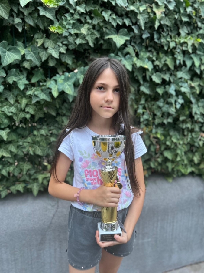 Ein junges Mädchen mit langen braunen Haaren steht im Freien vor grünem Efeu und hält eine goldene Trophäe in der Hand. Sie trägt ein T-Shirt mit Blumenmuster und graue Shorts.