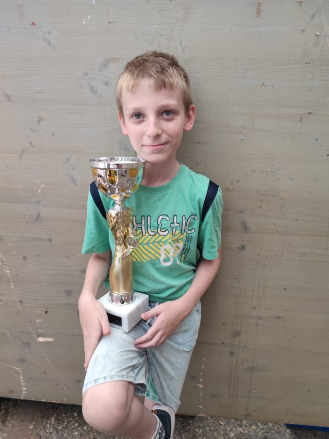 Ein kleiner Junge mit kurzen blonden Haaren, der ein grünes T-Shirt und helle Shorts trägt, sitzt vor einem schlichten Holzhintergrund, hält eine goldene Trophäe in der Hand und lächelt.