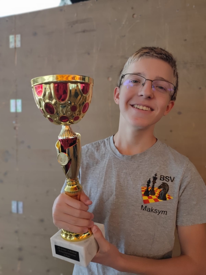 Ein lächelnder Teenager mit Brille und einem grauen BSV-Maksym-Shirt hält eine große goldene Trophäe mit roten Akzenten und einem weißen Sockel in den Händen und steht vor einer schlichten braunen Wand.