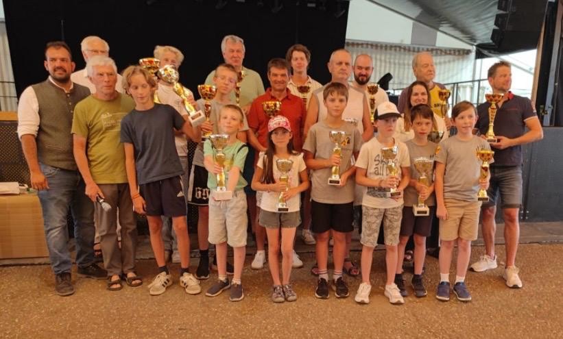 Eine Gruppe von Erwachsenen und Kindern steht in einem Raum, hält Pokale in den Händen und lächelt, was darauf hindeutet, dass sie bei einer Veranstaltung oder einem Wettbewerb gewonnen haben.