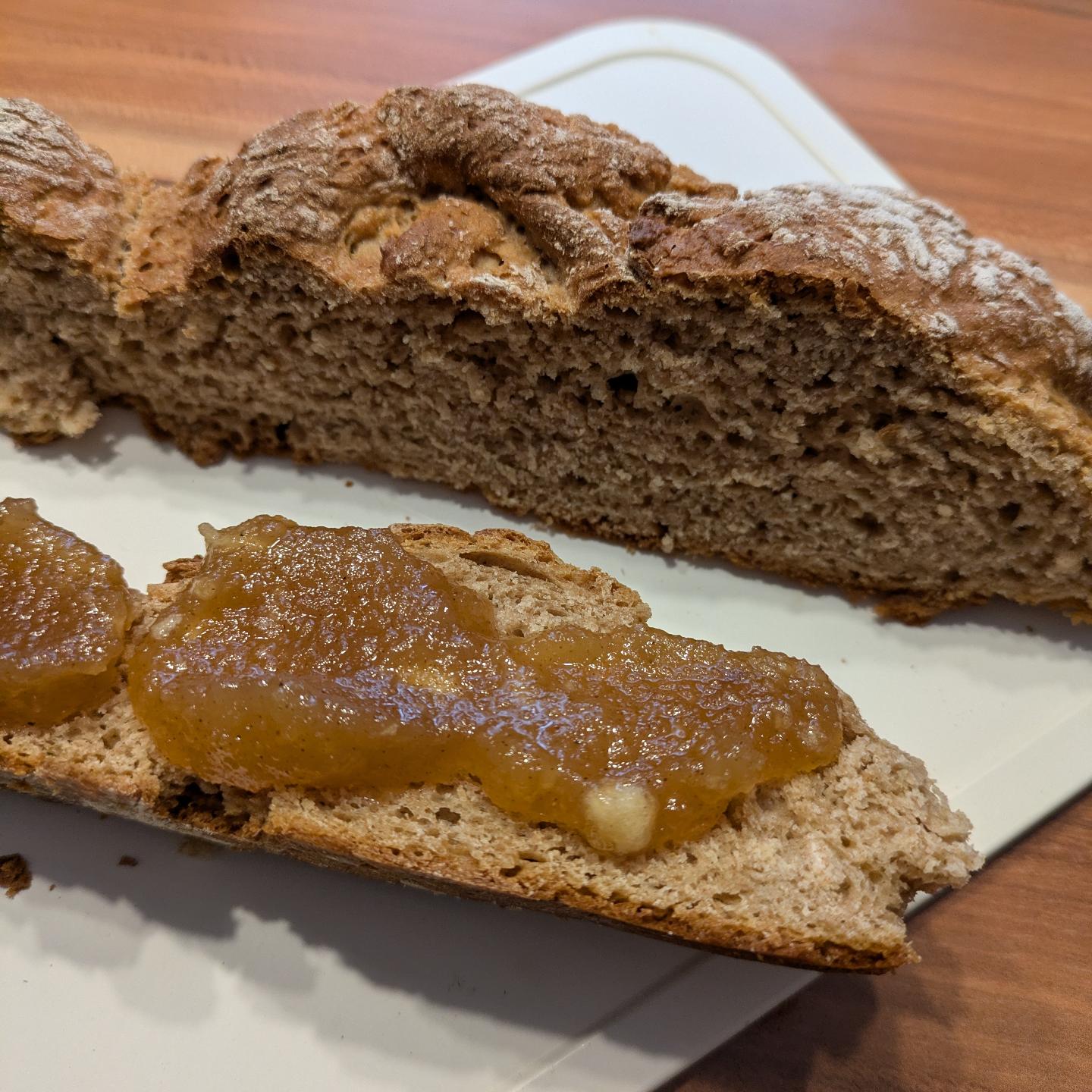 Eine Scheibe rustikales Schwarzbrot mit einer großzügigen Schicht Apfelmarmelade liegt auf einem weißen Schneidebrett, dahinter eine weitere einfache Scheibe Brot. Die Oberfläche darunter ist eine hölzerne Arbeitsplatte.