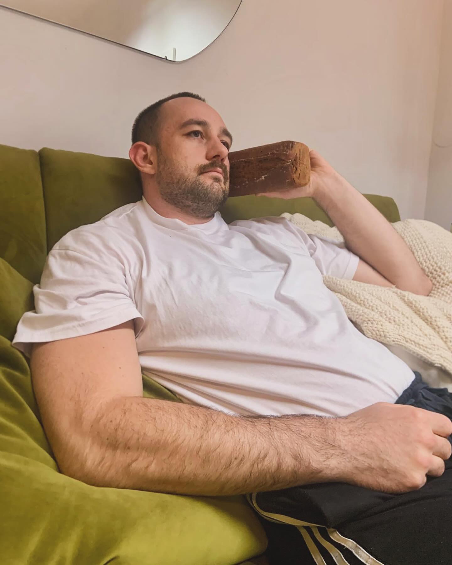 Ein Mann in einem weißen T-Shirt sitzt auf einer grünen Couch und hält sich einen Laib Brot ans Ohr, als ob er damit telefonieren würde. Er sieht nachdenklich aus, neben ihm liegt eine leichte Decke.