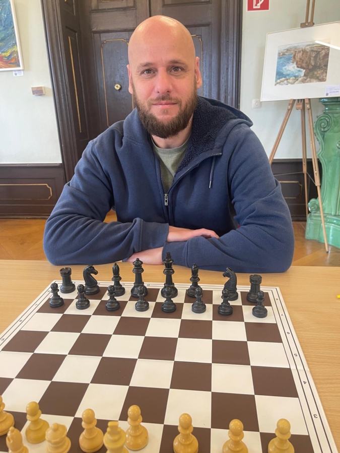 Ein Mann mit Bart und marineblauer Jacke sitzt am Schachbrett, die Arme verschränkt, bereit zu spielen. Die Schachfiguren sind für ein neues Spiel aufgestellt. Im Hintergrund sind Kunstwerke und eine Holztür zu sehen.