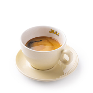 Eine Tasse Espresso mit einer cremigen Cremaschicht, serviert in einer hellbeigen Tasse mit passender Untertasse. Auf dem Innenrand der Tasse steht in gelber Schrift das Wort "Strich". Der Hintergrund ist einfarbig weiß.