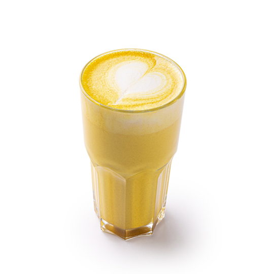 Ein Glas goldener Kurkuma-Latte mit einem herzförmigen Muster im Schaum vor einem weißen Hintergrund.