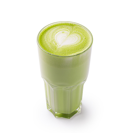 Ein klares Glas, gefüllt mit einem cremig-grünen Matcha-Latte, gekrönt von einem herzförmigen Latte-Art-Motiv aus weißem Schaum, vor weißem Hintergrund.