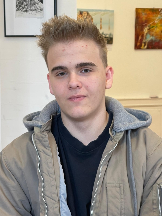 Eine junge Person mit kurzem, hellbraunem Haar und neutralem Gesichtsausdruck trägt eine hellbraune Jacke mit grauer Kapuze über einem schwarzen Hemd und sitzt in einem Innenraum mit Kunstwerken an den weißen Wänden hinter ihnen.