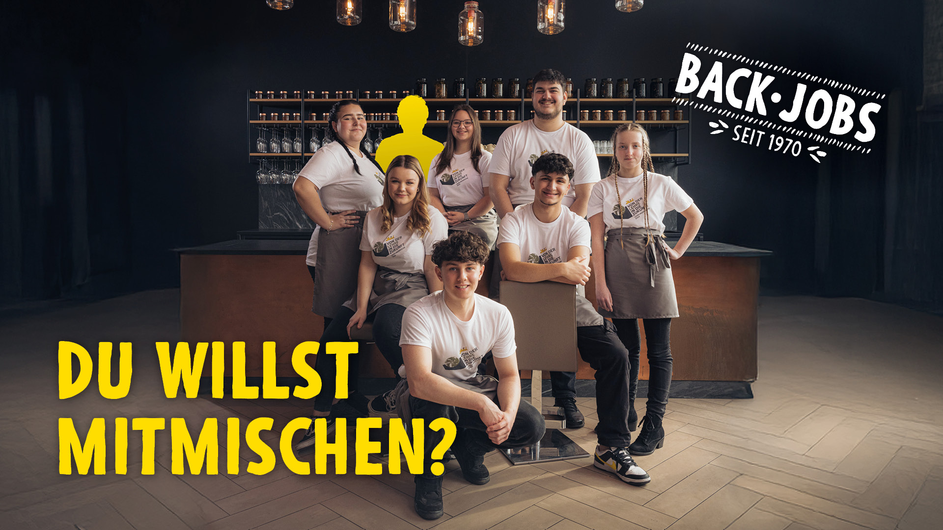 Sieben junge Leute in weißen Hemden und Schürzen posieren zusammen in einer Bäckerei. In großer gelber Schrift steht "Du willst mitmischen?" und auf einem Schild steht "Back-Jobs seit 1970". Eine Silhouette ist gelb hervorgehoben.